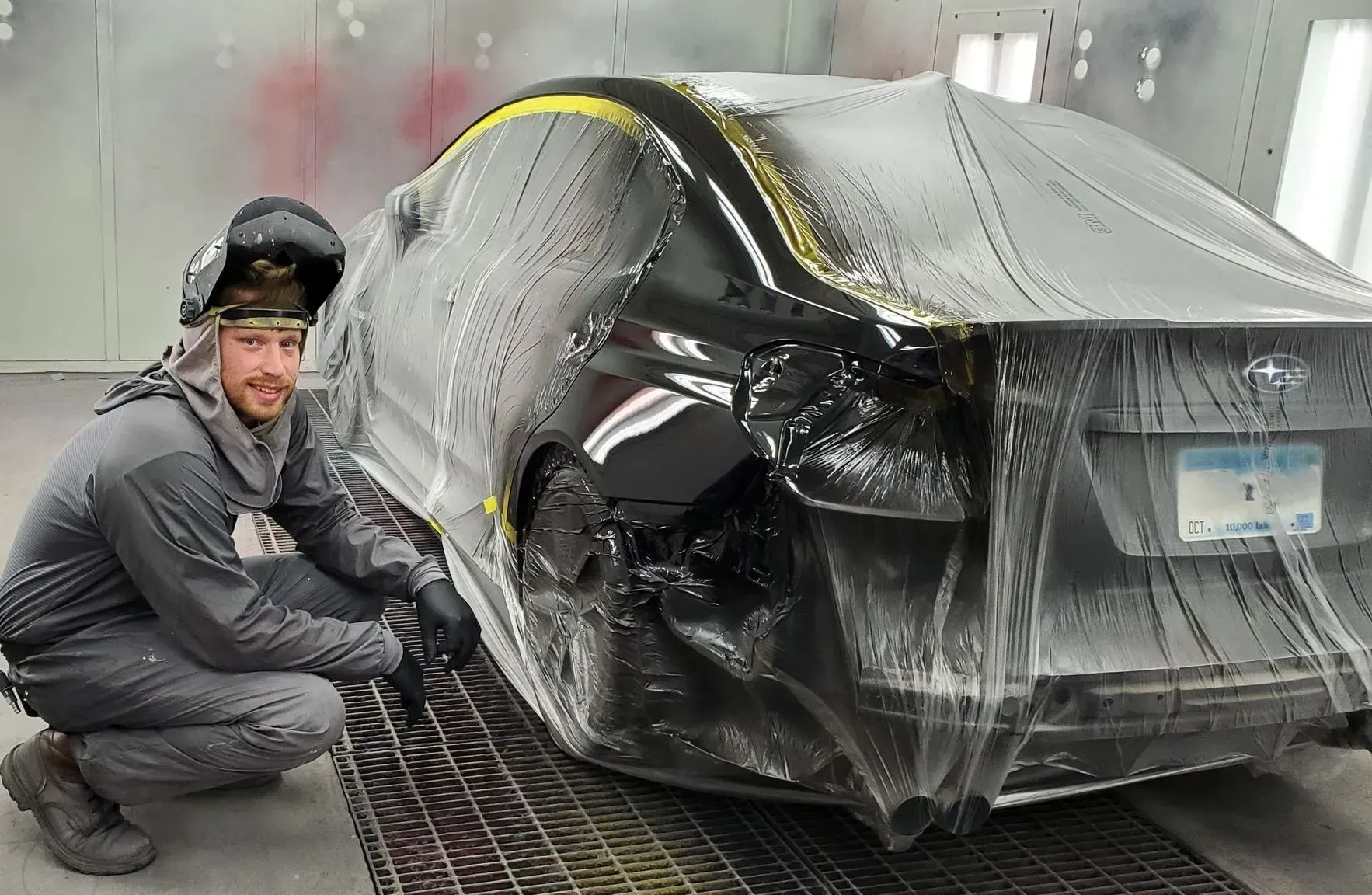 Worker crouches beside a black car wrapped in clear protective plastic in a paint booth