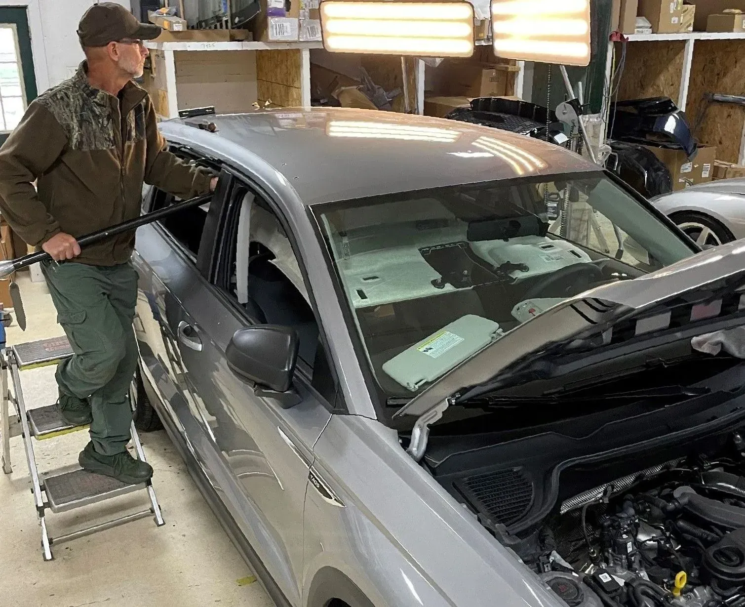 Mechanic inspecting a silver car in a garage with the hood open and bright work lights overhead