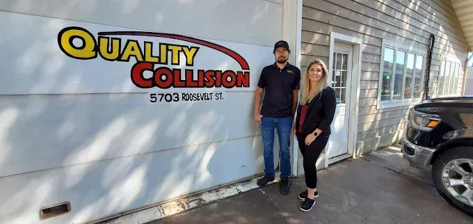 Two people standing outside Quality Collision auto body shop beside a white building.