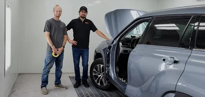 Two men stand beside a gray car in a garage, one holding a yellow tool near the front wheel.