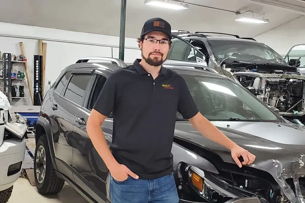 Man standing beside a gray SUV in an auto shop, with a black car parked behind him.