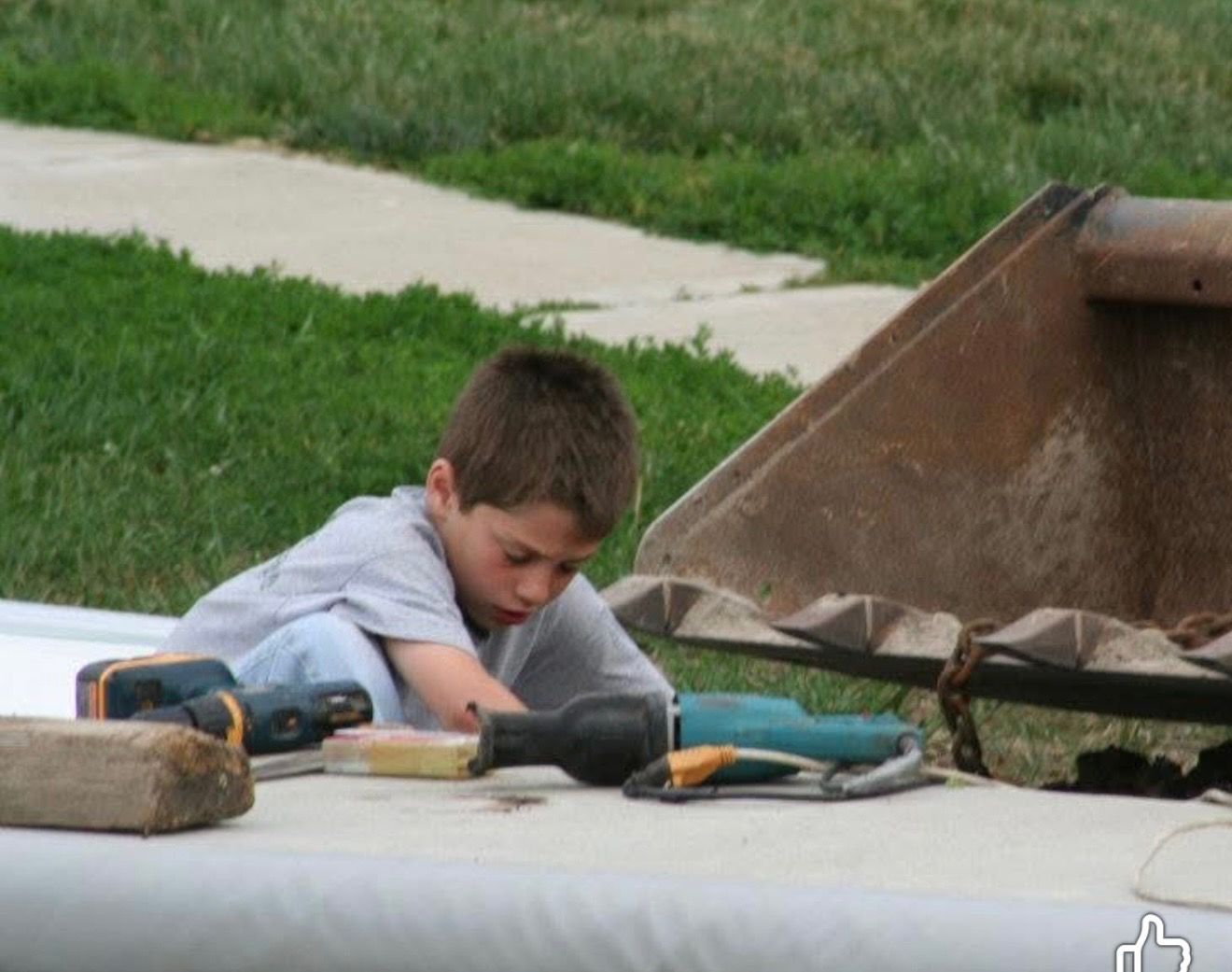 Boy playing with toy construction tools beside a large wheelbarrow on a sidewalk, with grass and a path nearby
