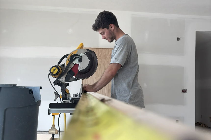 Man using a miter saw in a room under construction, cutting wood with a tape measure in the foreground