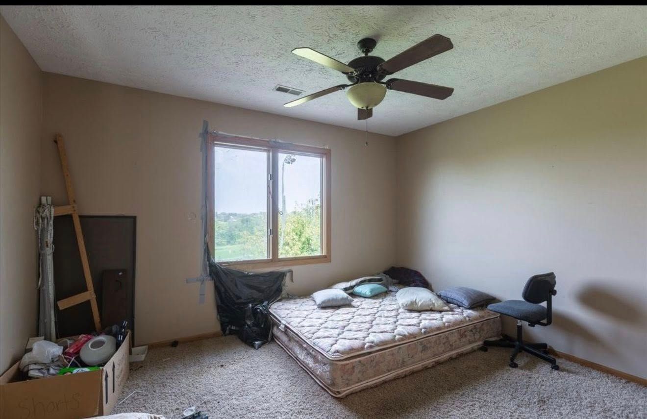 Spare bedroom with a bed, office chair, window, ceiling fan, and a ladder leaning against the wall.