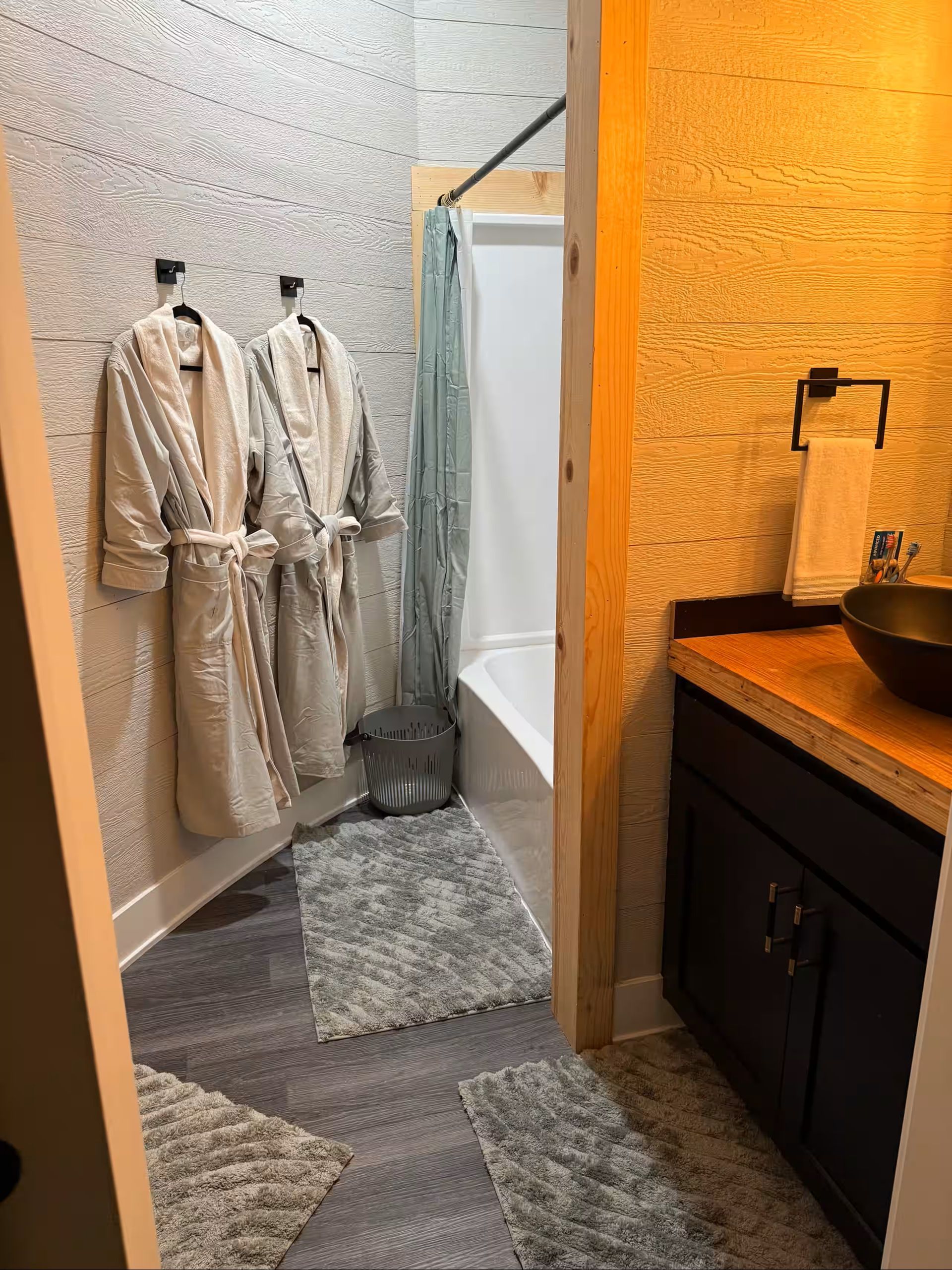 Small dressing area with two beige robes on hooks, a mirror, rug, and a sink counter in warm light.