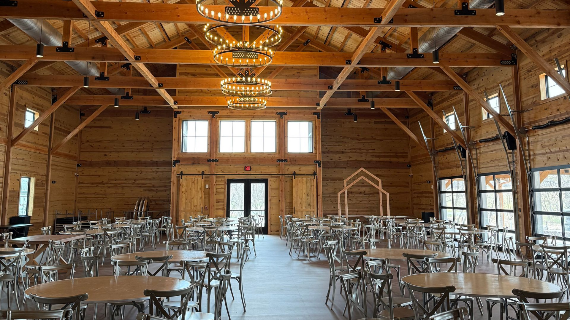 Empty rustic banquet hall with wooden beams, chandeliers, and rows of tables and chairs