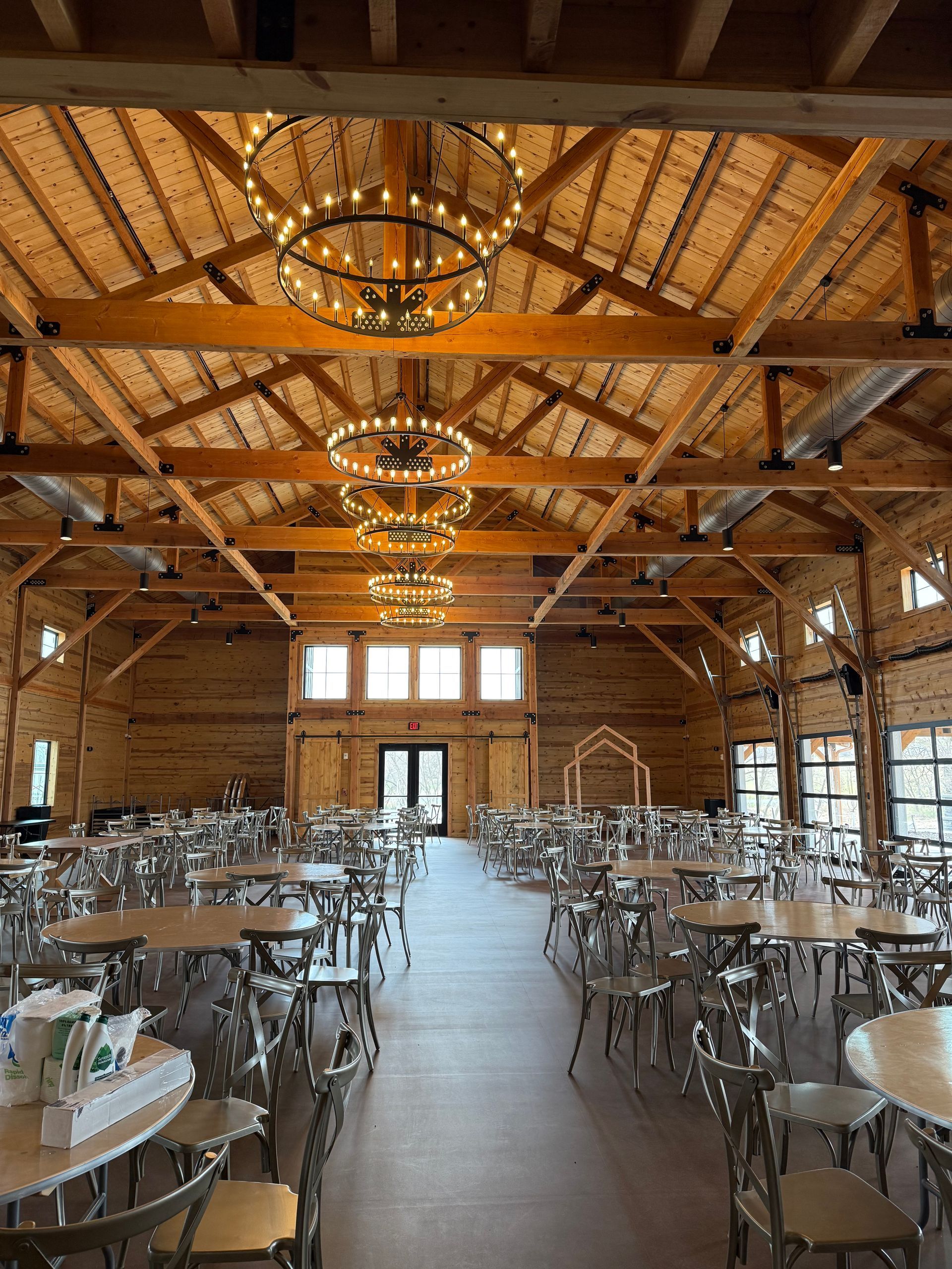 Rustic banquet hall with wooden beams, chandeliers, and rows of silver chairs and tables