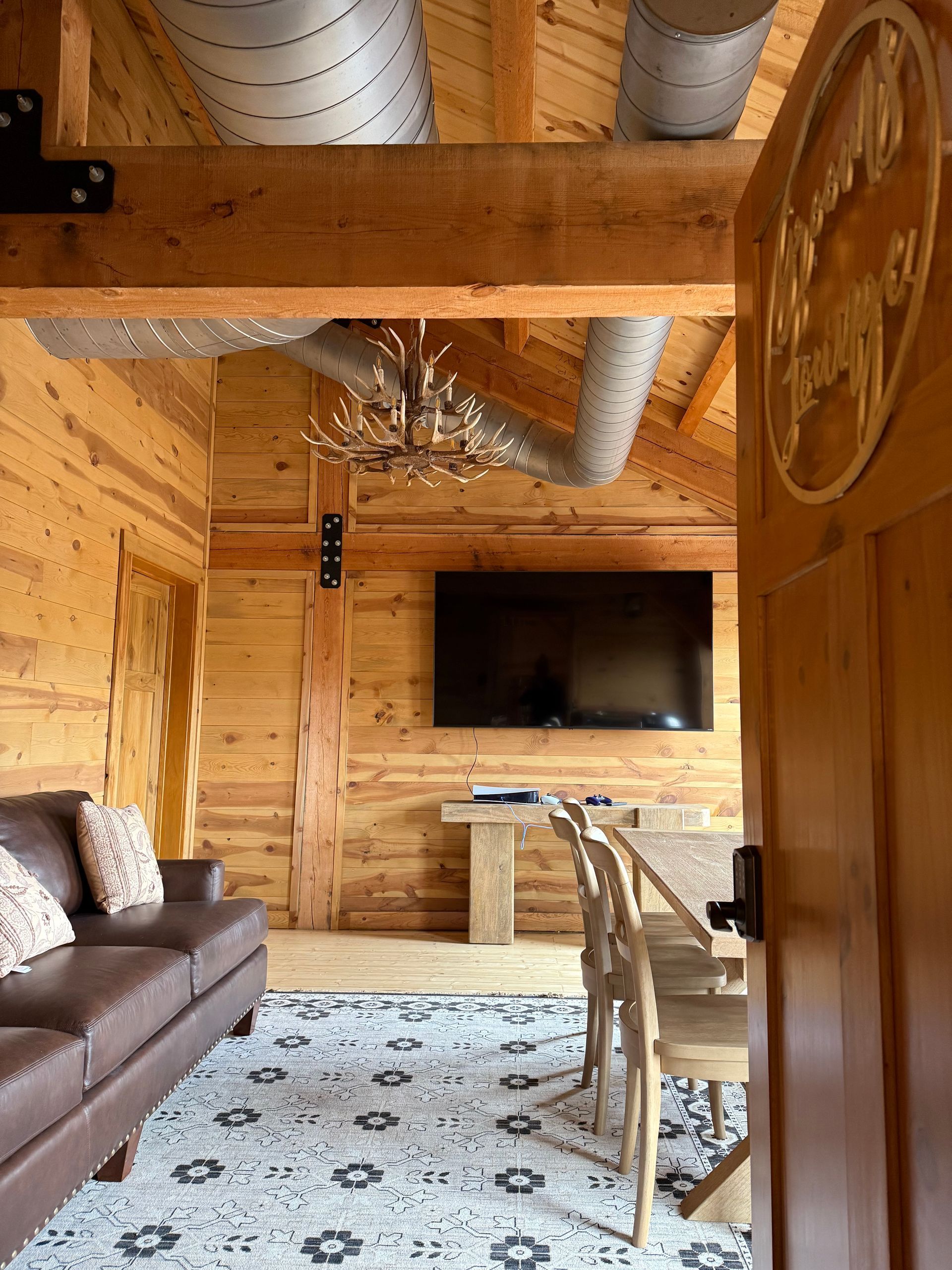 Wood-paneled cabin living room with brown sofa, dining table, wall TV, and white patterned rug