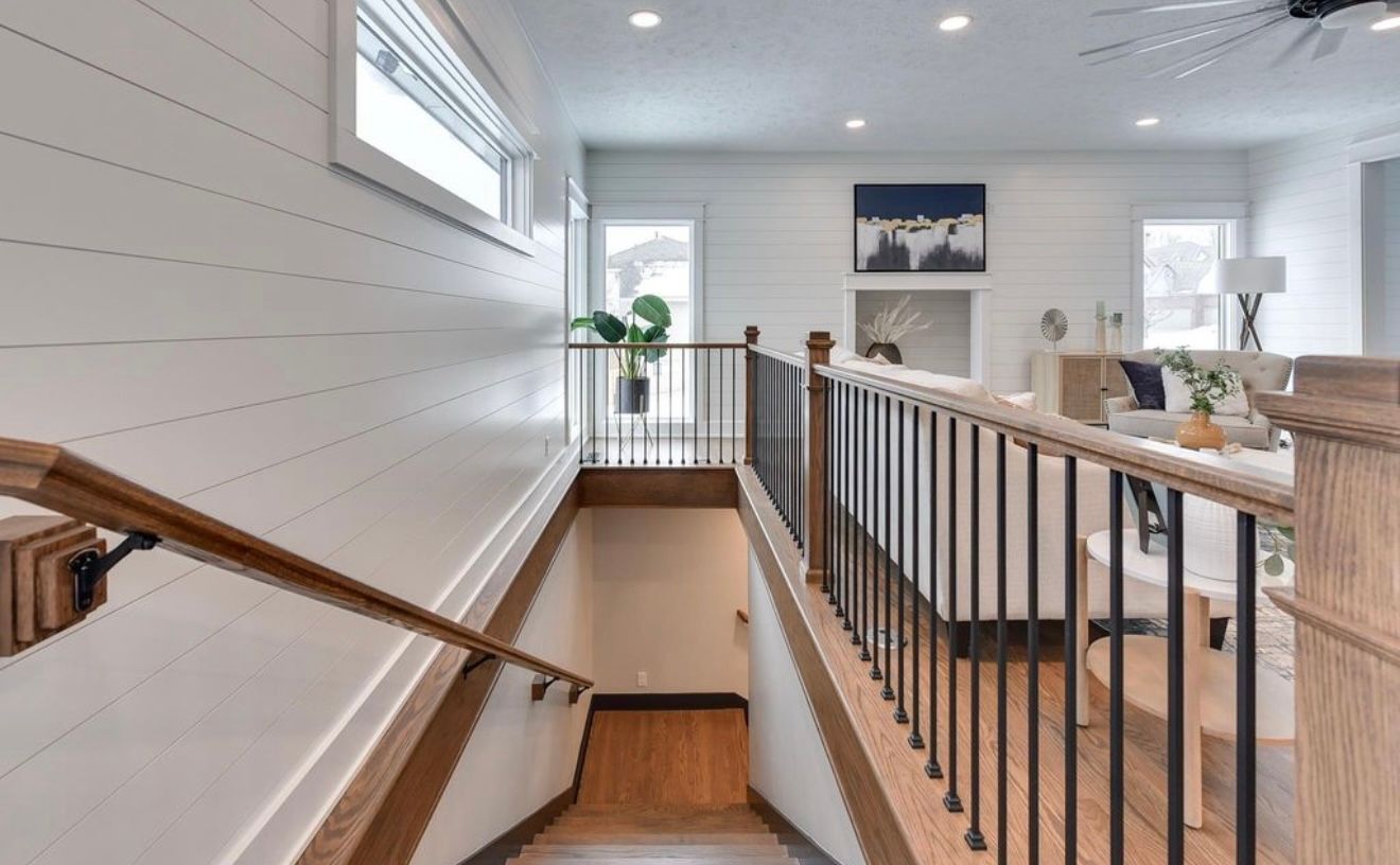 Bright upstairs hallway with wooden railing, staircase, white walls, and large windows looking into a modern home