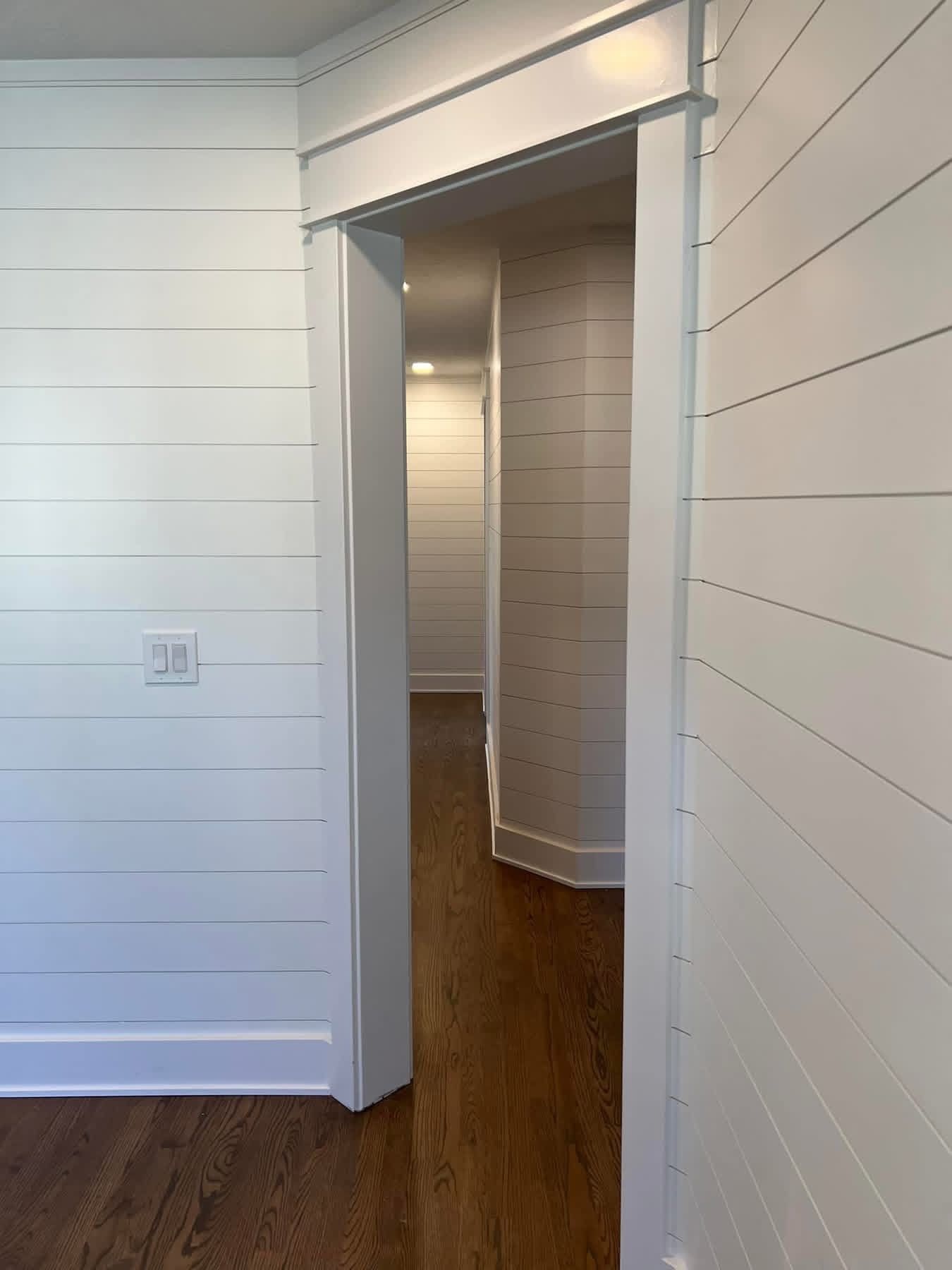 White hallway with shiplap walls, wood floor, and open doorway leading to a narrow corridor.