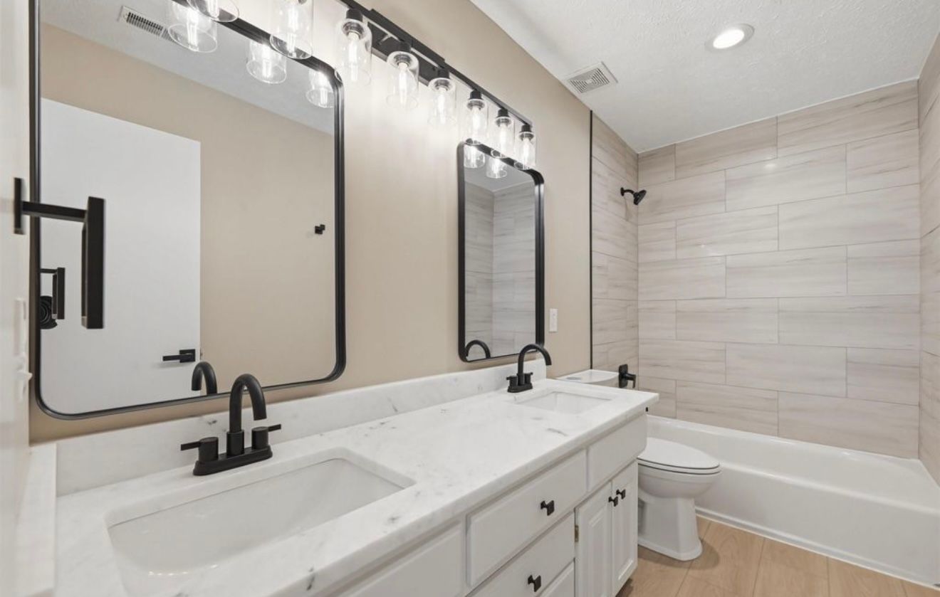 Bright modern bathroom with double vanity, large mirrors, black faucets, and a tub with tiled shower wall