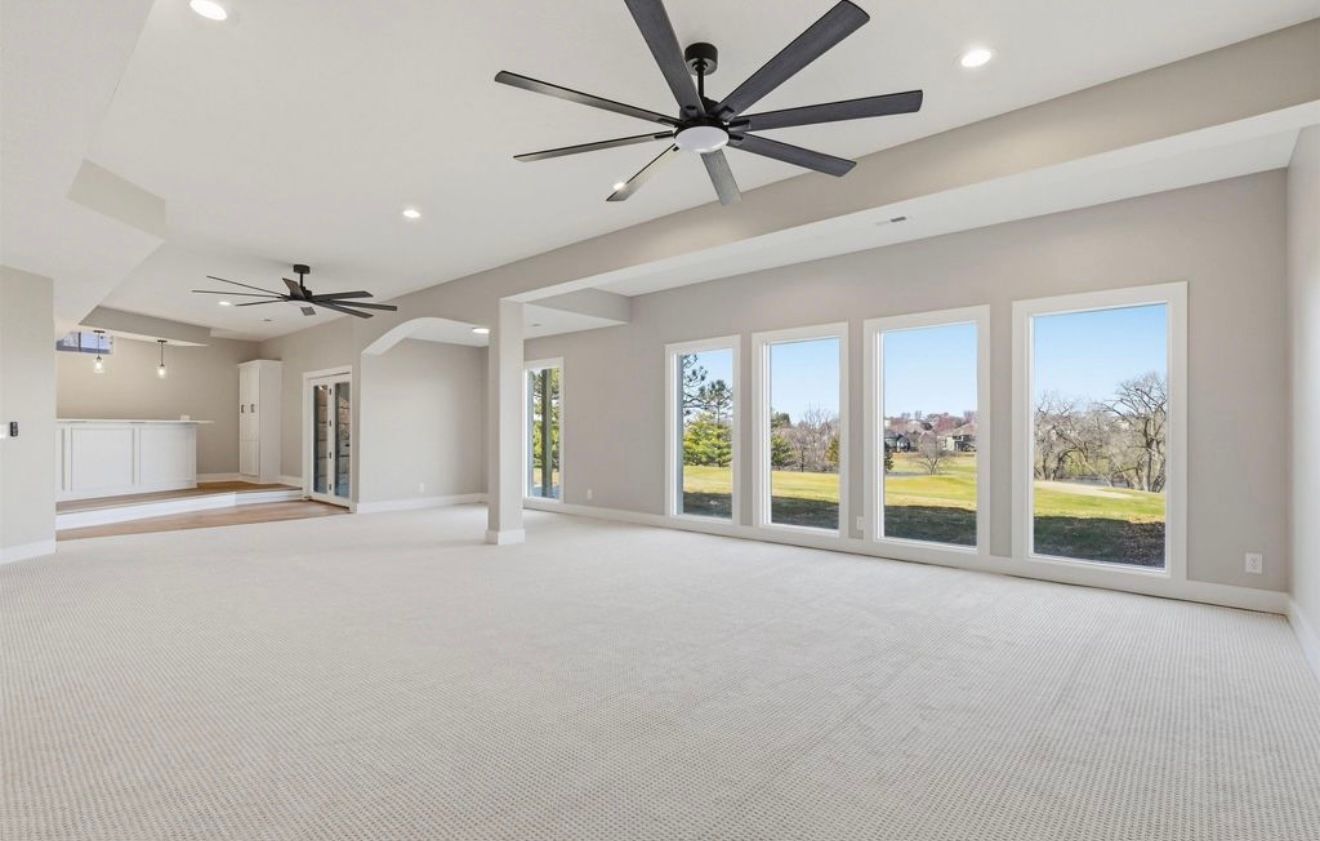 Spacious empty living room with carpet, ceiling fans, and large windows overlooking a green backyard