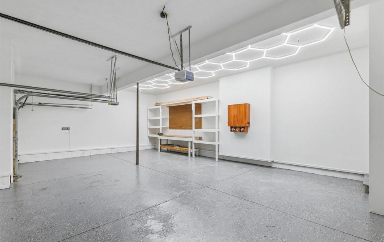Empty garage with gray floor, white walls, workbench, overhead lighting, and an orange wall cabinet