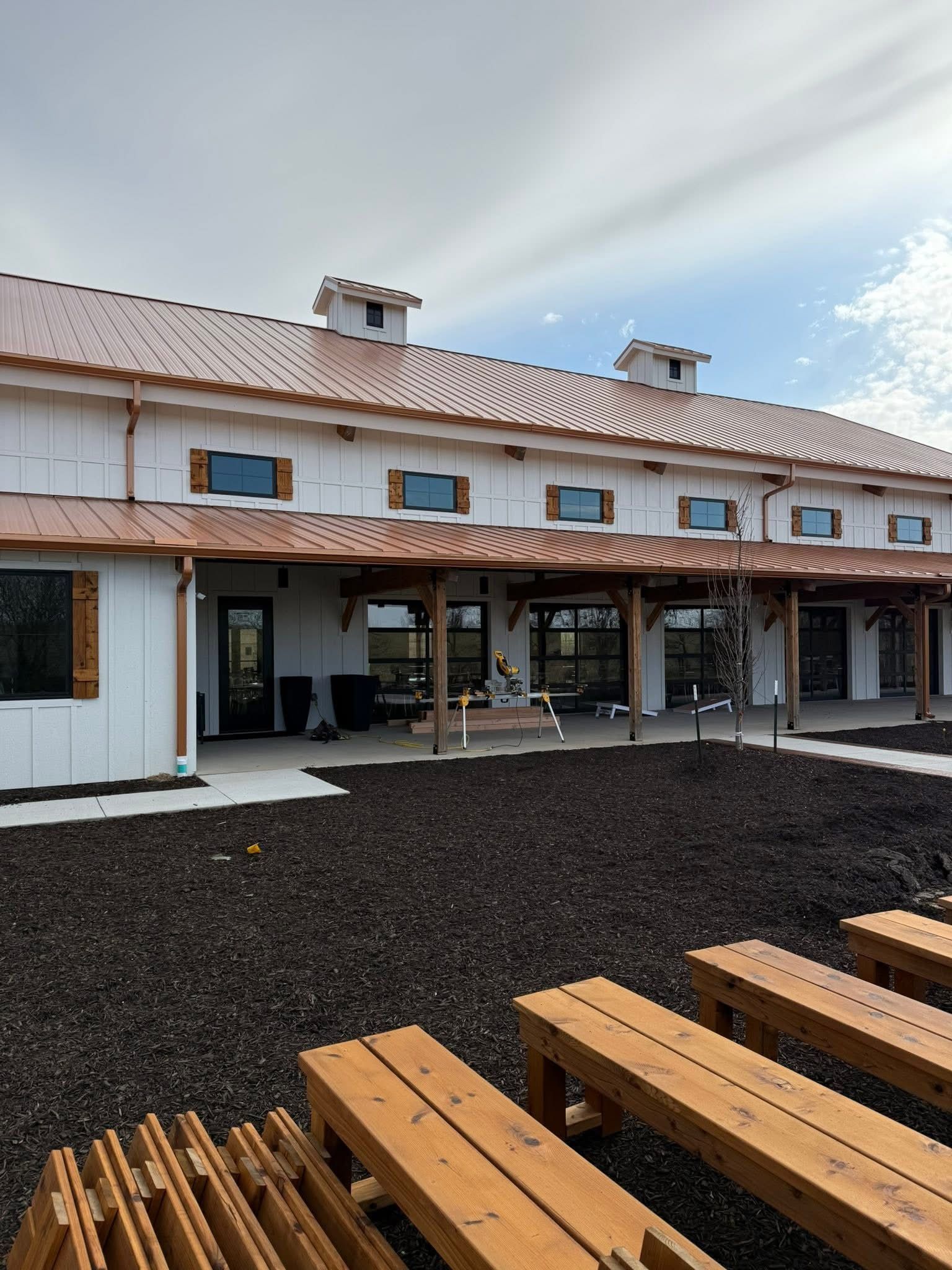 Patio with wooden picnic tables facing a white stucco building with terracotta roof and shaded porch.