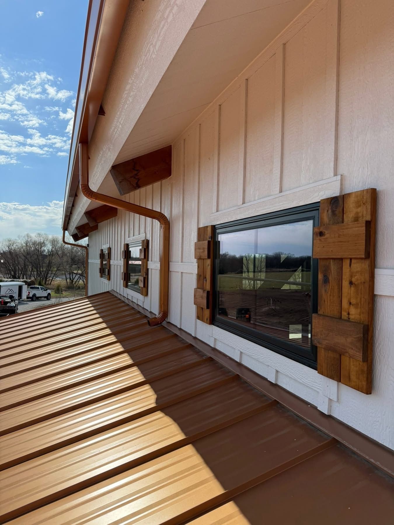 Exterior house wall with windows and brown metal roof, viewed from a sunlit balcony or walkway.