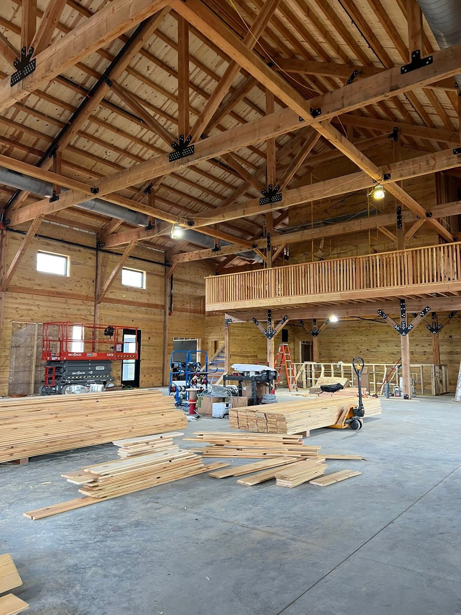 Wooden timber-frame barn interior with stacked lumber, scissor lifts, and exposed beams under construction