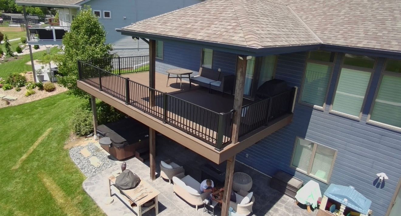 Covered backyard deck with grill, railing, patio seating, and outdoor stairs beside a blue house.