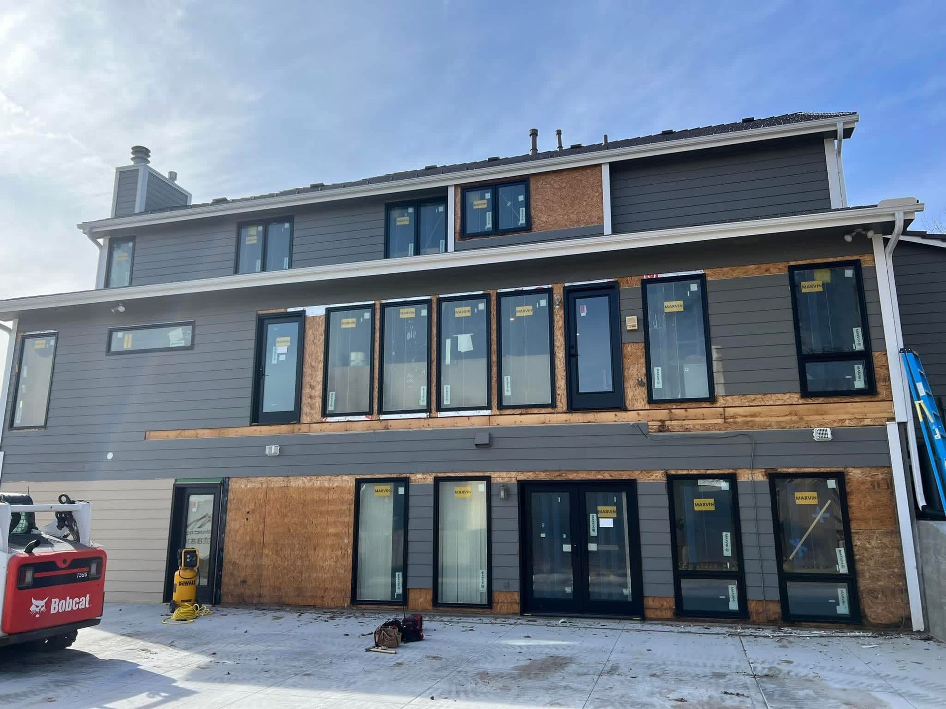Modern gray townhouse exterior with large windows, wooden balcony, and construction equipment in front