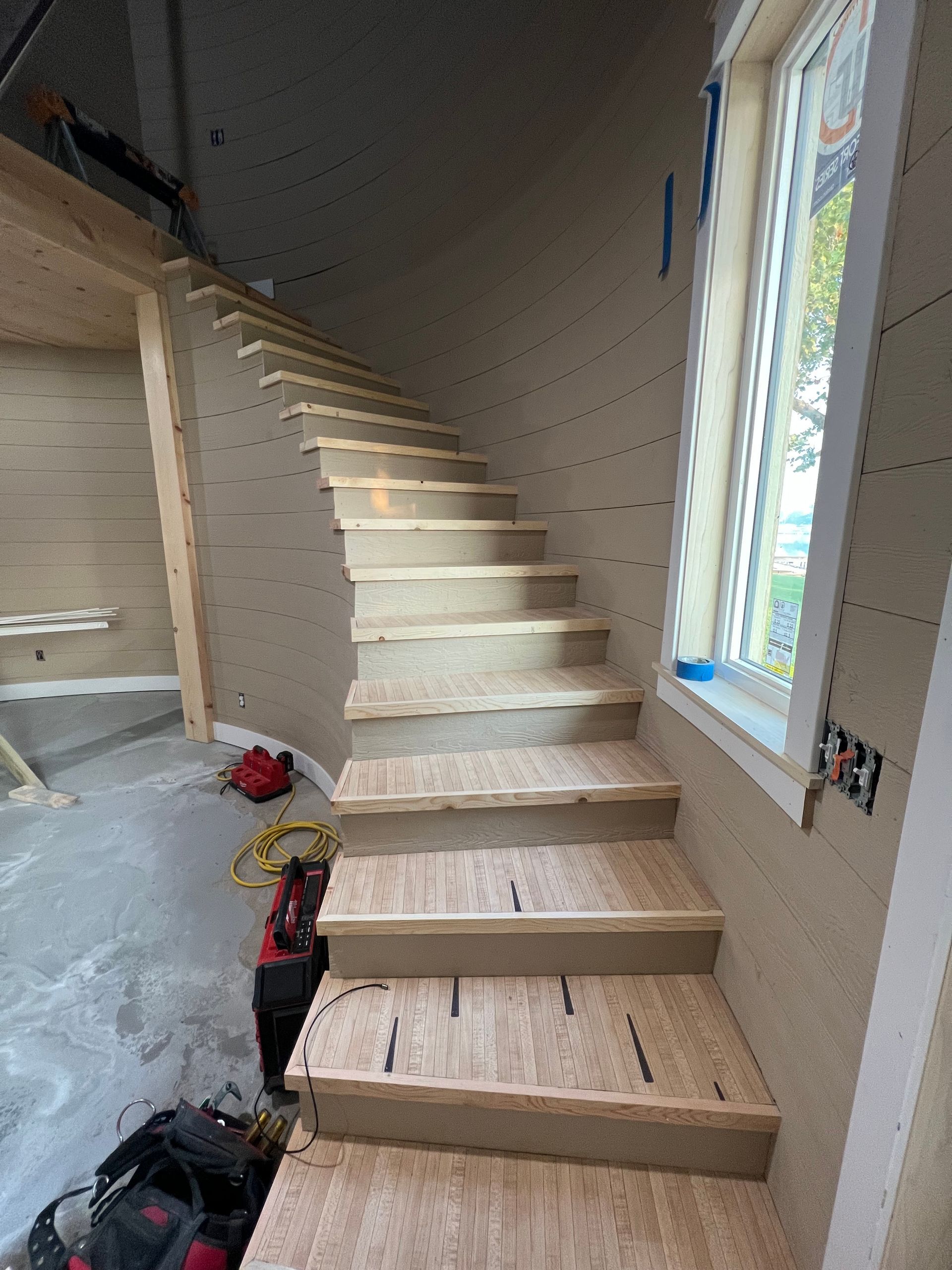 Unfinished wooden staircase in a home interior beside a tall window.