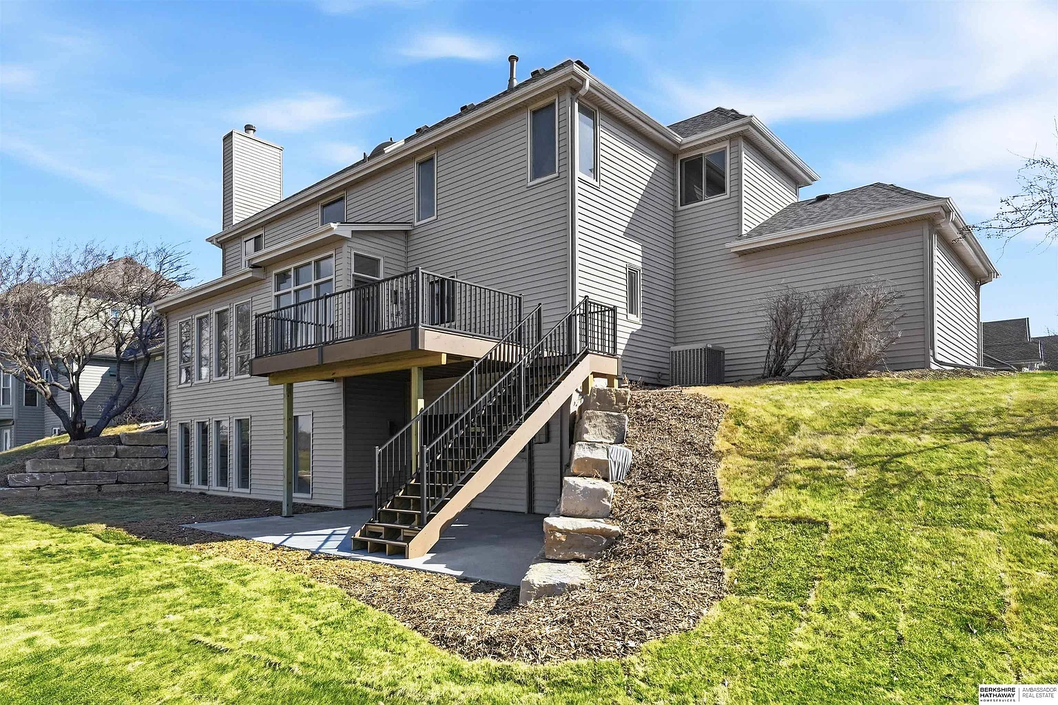 Two-story beige house with wood deck, exterior stairs, and stone retaining wall on a sunny hillside