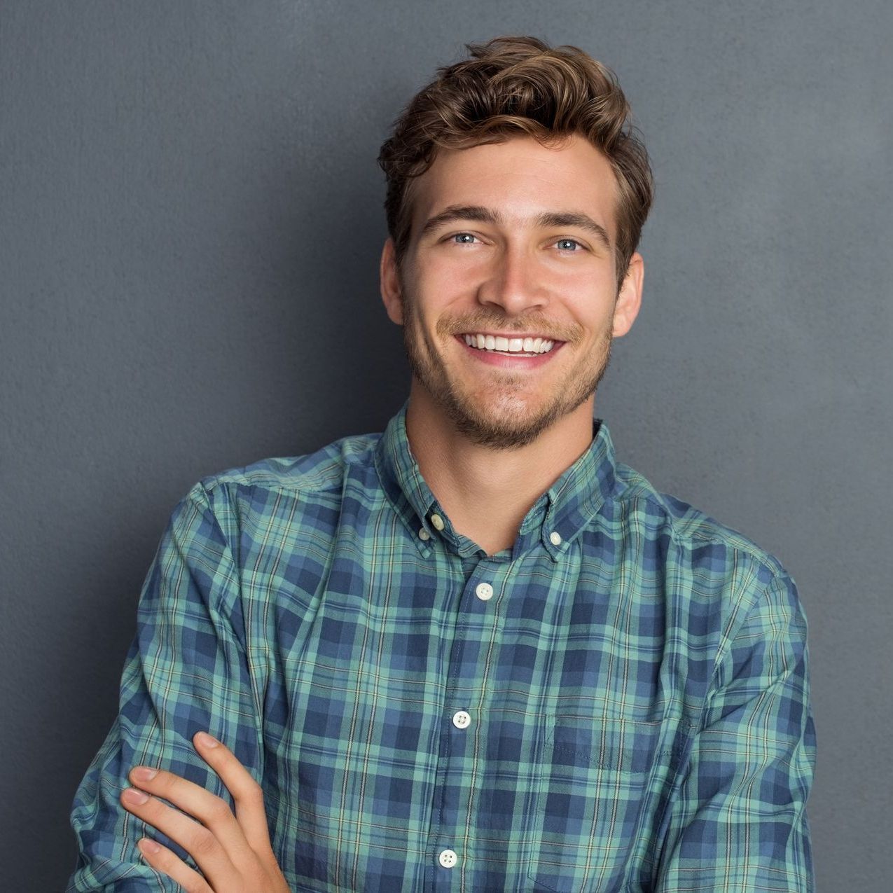 Man with curly brown hair smiles, arms crossed, wearing a blue and green plaid shirt in front of a grey background.