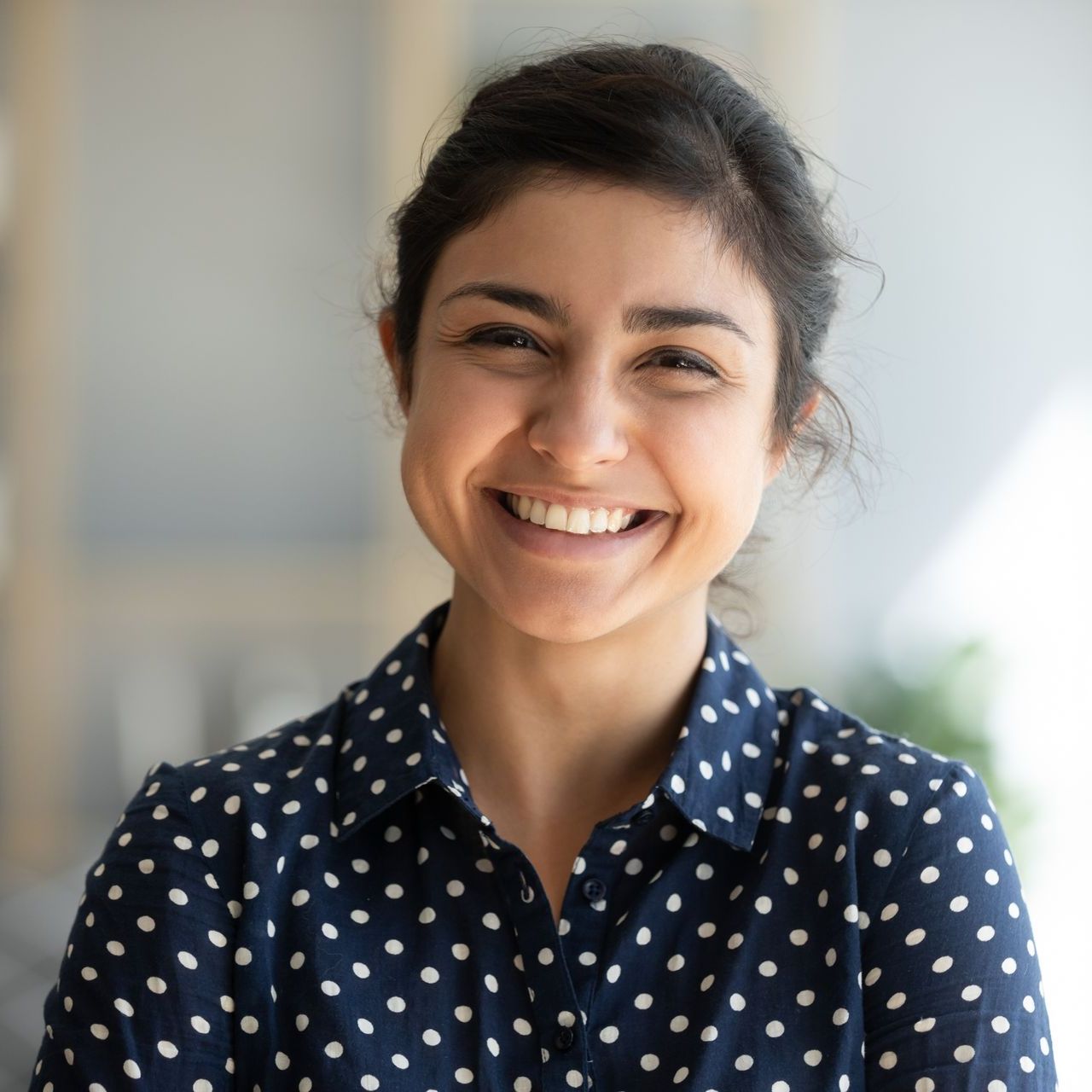 Woman with dark hair pulled back smiles broadly, wearing a blue polka-dot shirt. Soft lighting.