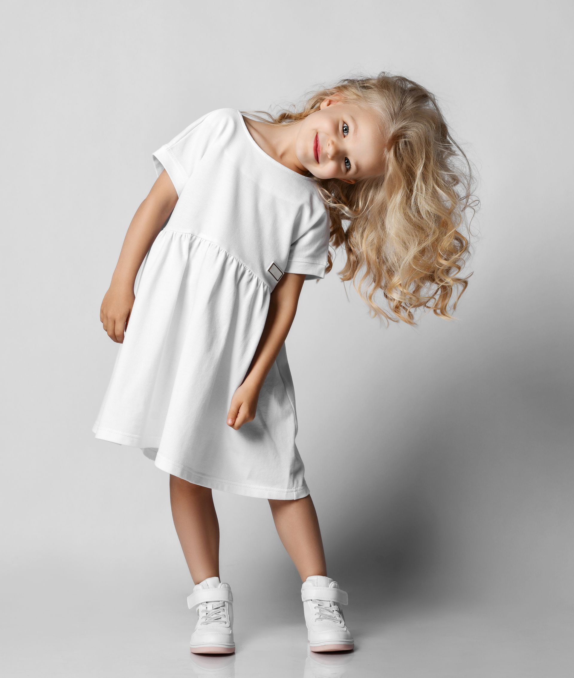 A little girl in a white dress and white sneakers is posing for a picture.