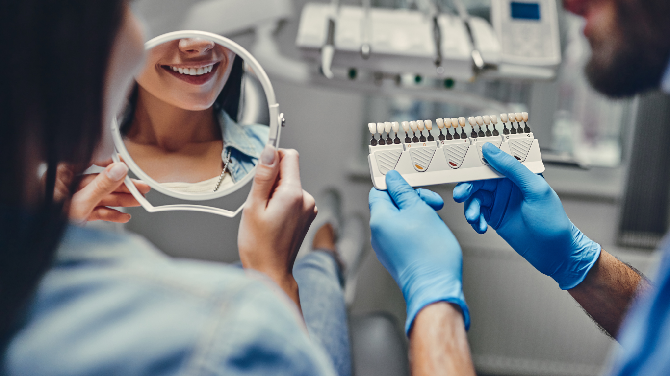 Dentist with shade guide consulting with patient, who is holding a mirror, in dental office.