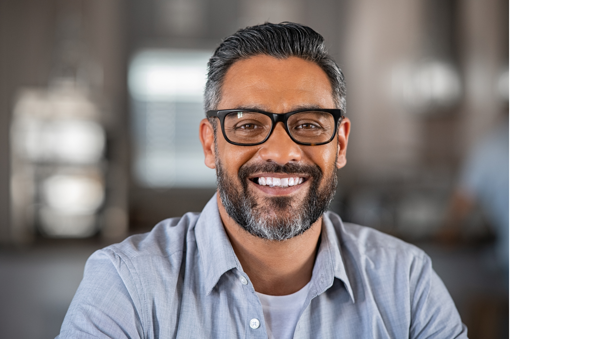 Man with glasses smiles, looking directly at the camera, wearing a light blue button-down shirt.