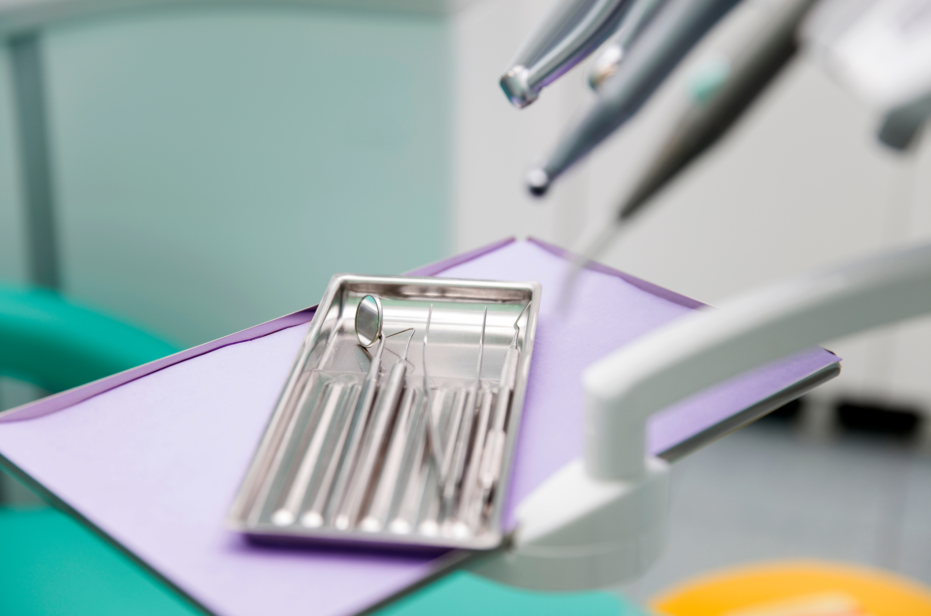 Dental tools in a metal tray on a purple drape, with dental chair and equipment in a clinic setting.