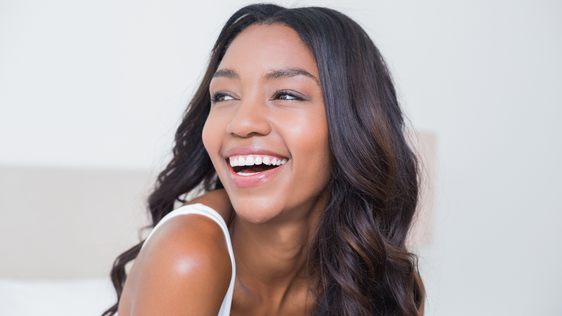 Woman with long wavy dark hair smiles broadly, looking to her right in a bright room.
