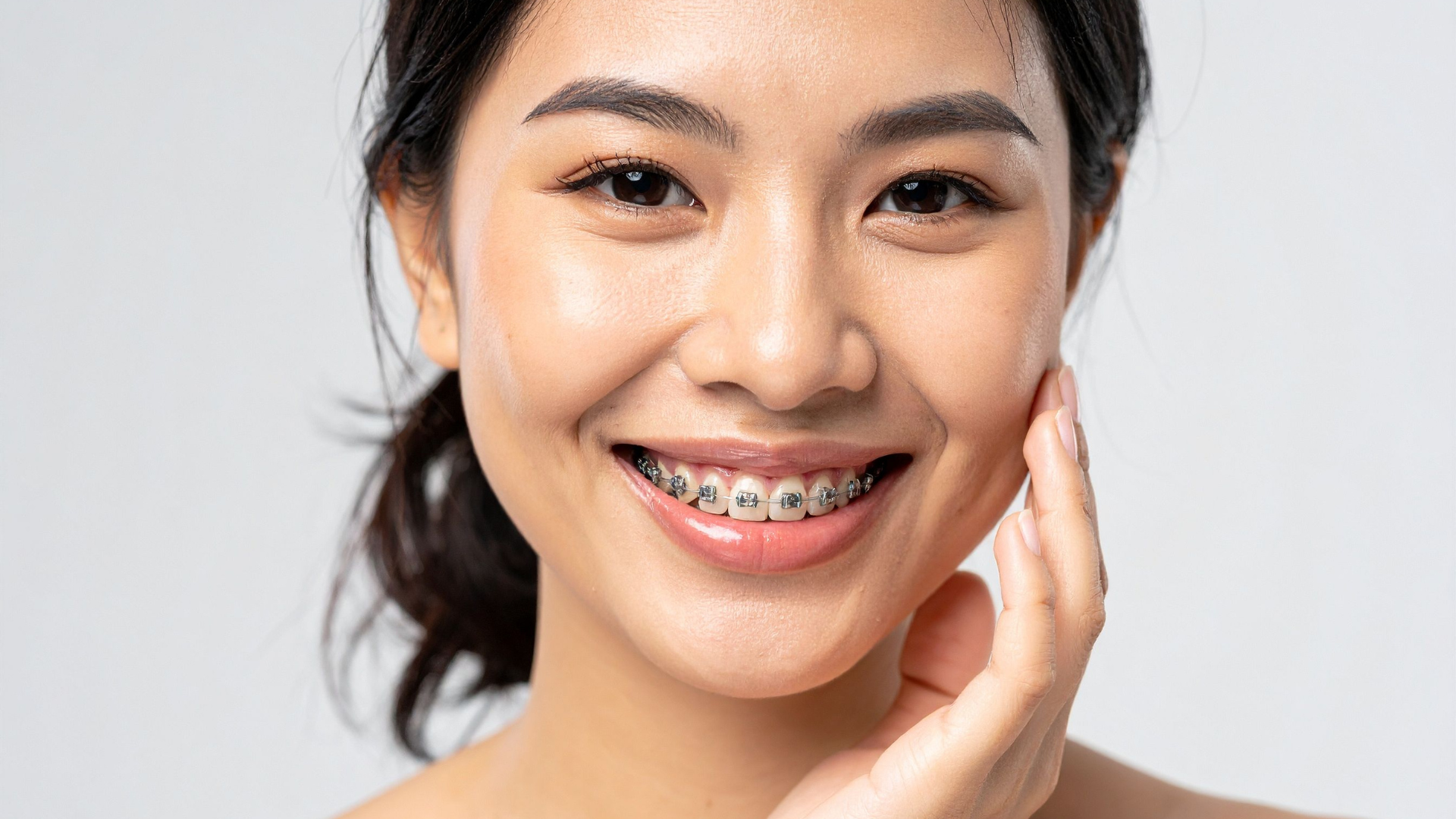 Woman with braces smiles, touching her cheek; bright, white background.