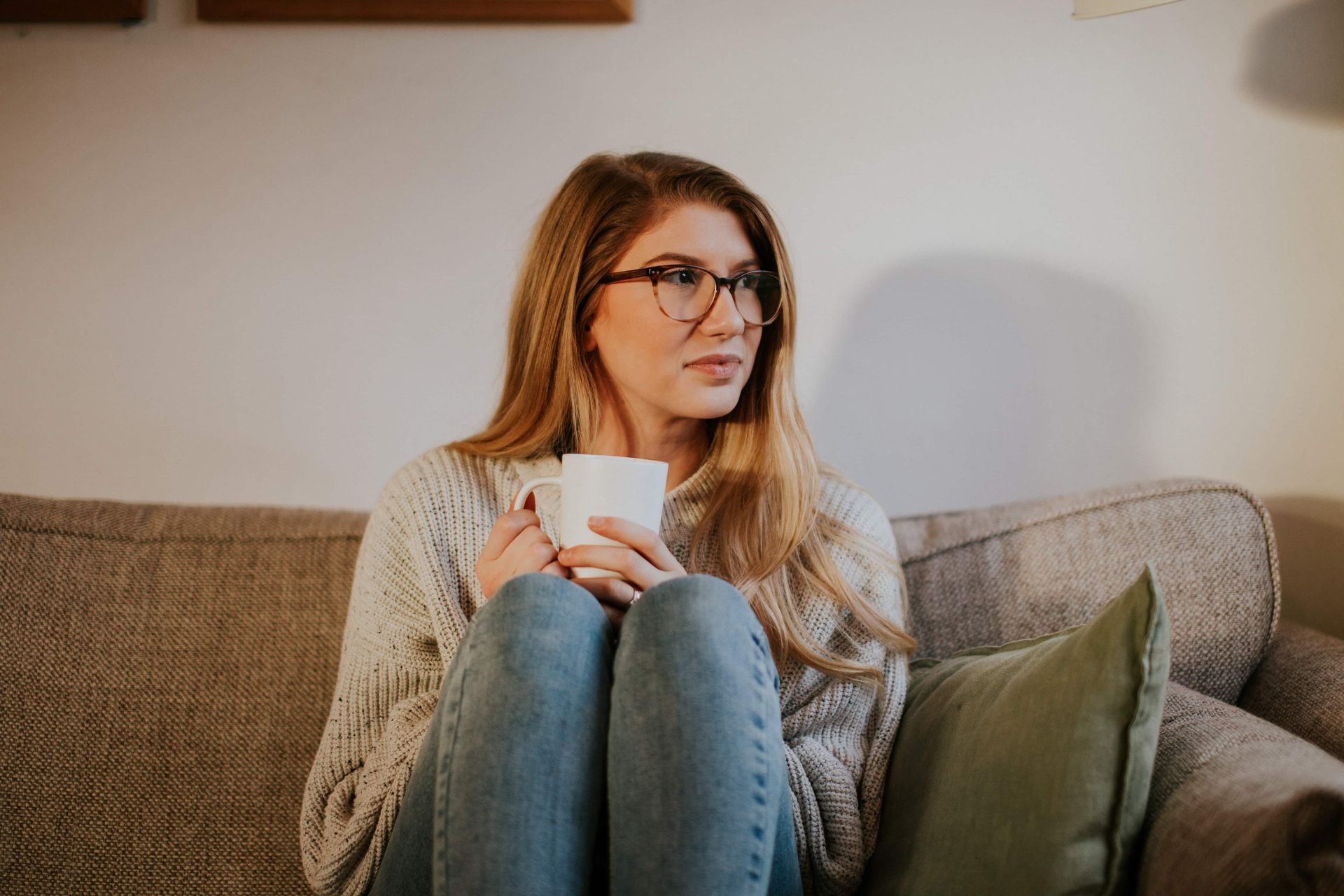 A person wearing glasses and a knit sweater sits on a couch, holding a white mug and looking toward the side.