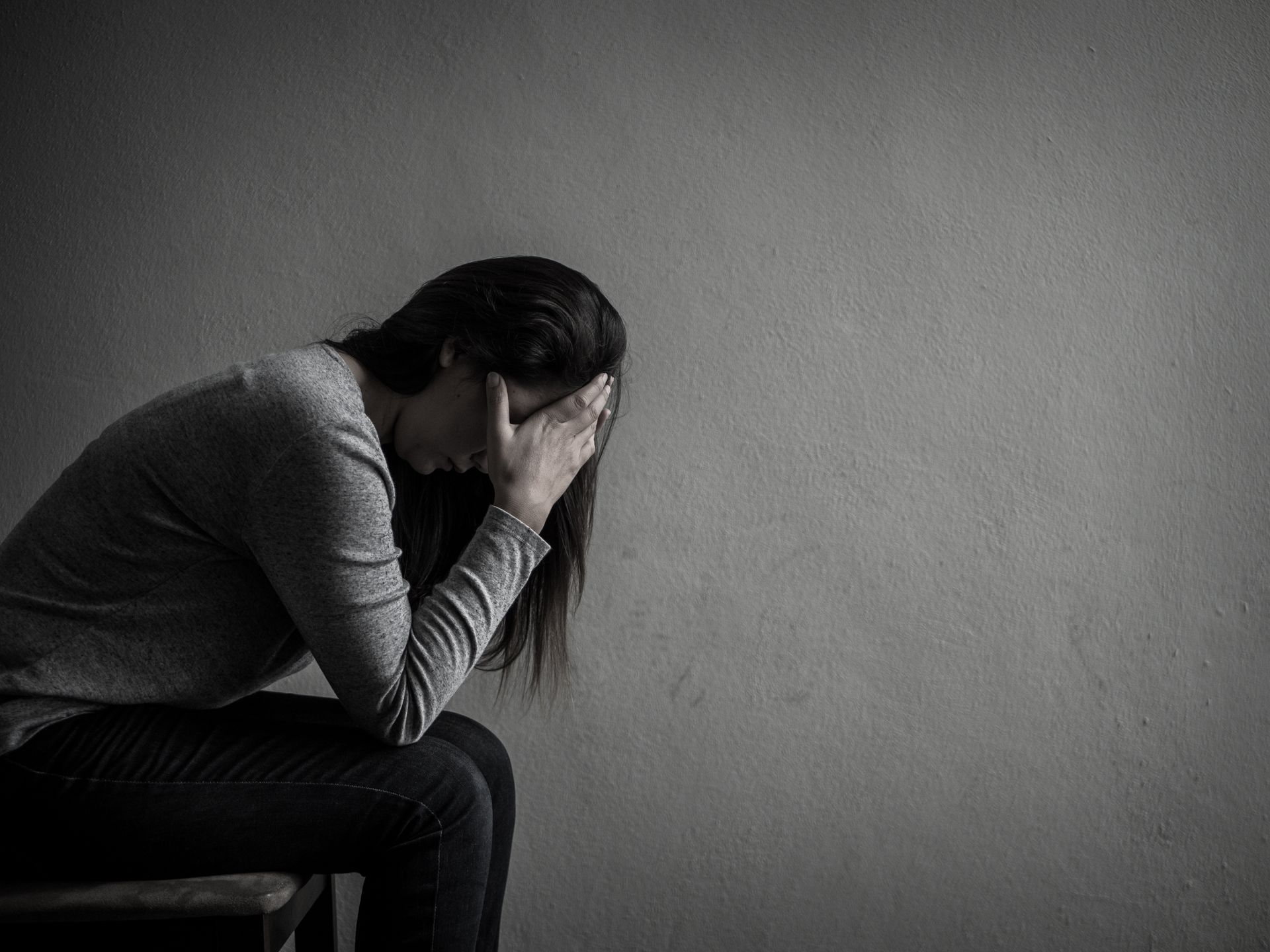 Person sitting on a bench, hunched over with face in hands against a plain gray wall.