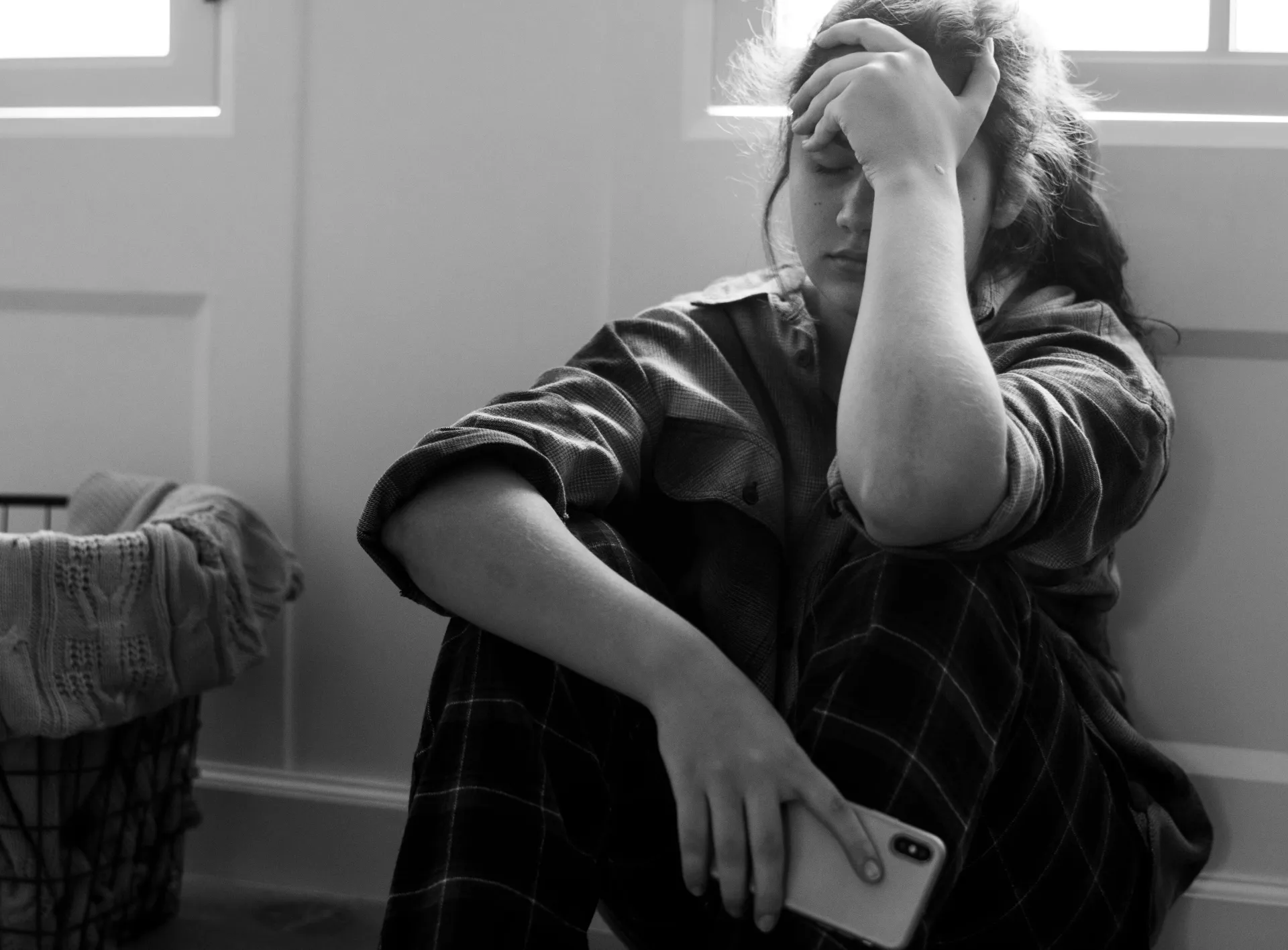 Person sitting on the floor by a window, head in hand, holding a phone, looking tired or upset