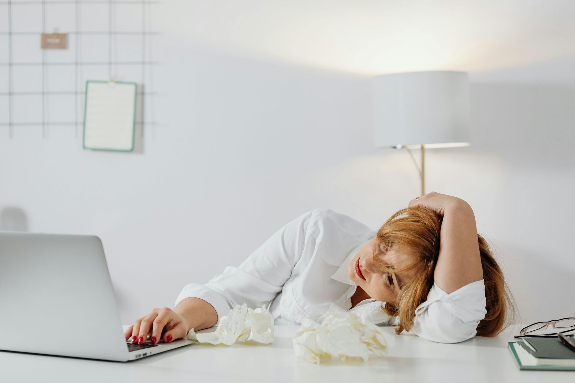 Sleepy person slumped at a desk with a laptop, hand on head, surrounded by scattered tissues.