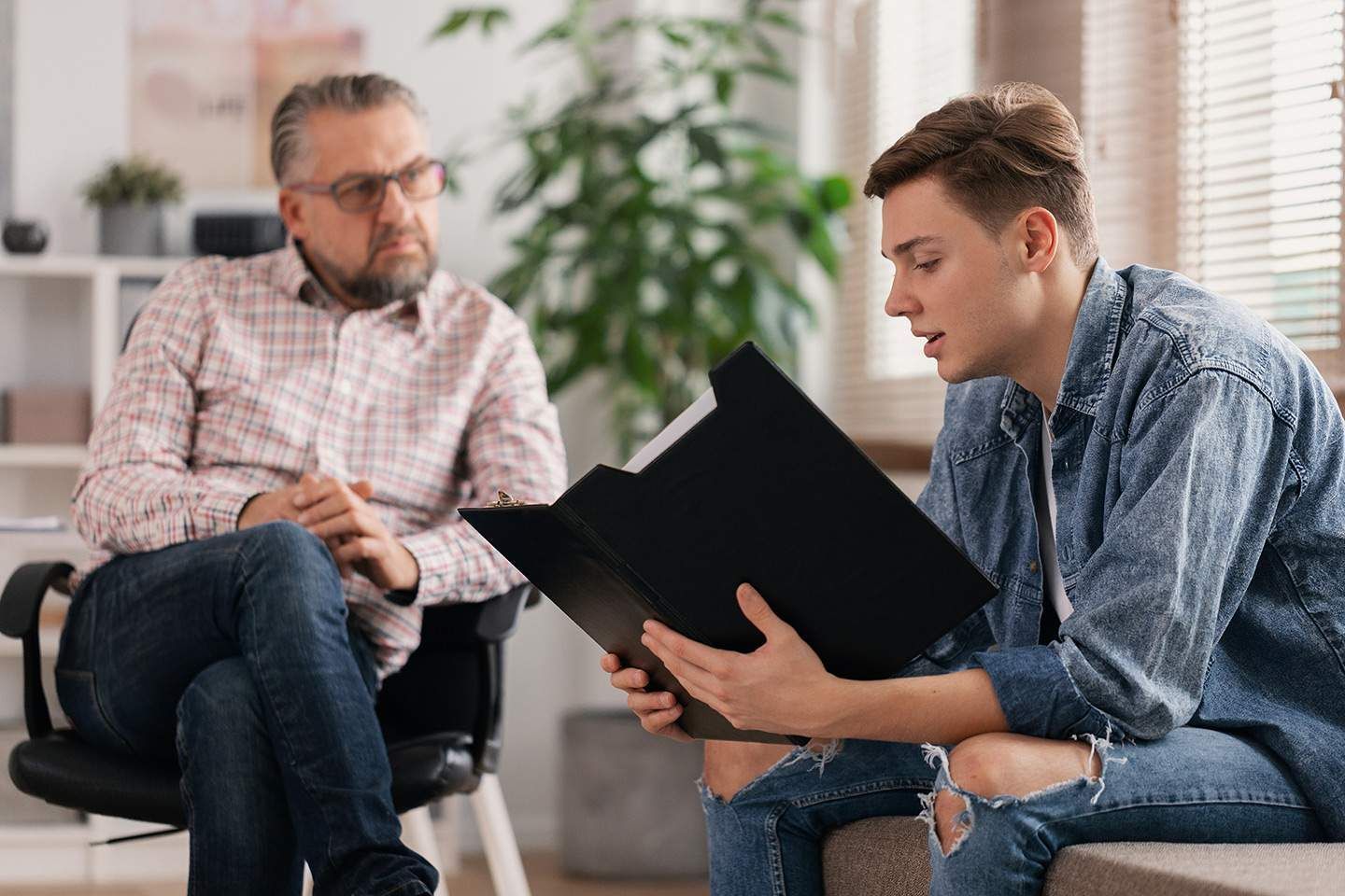 Teenager reading from a folder to a man seated in an office; both are looking at the folder.