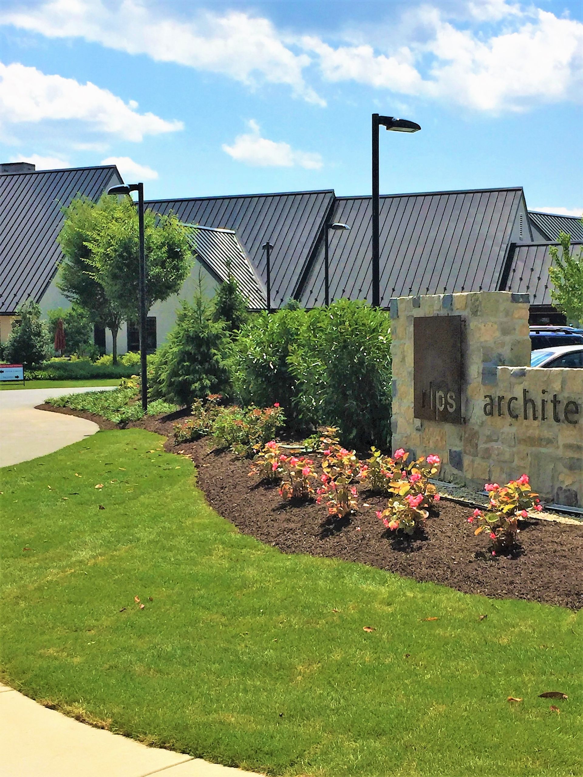 Landscaped lawn with flowers and shrubs beside a stone sign, in front of a large building under a blue sky