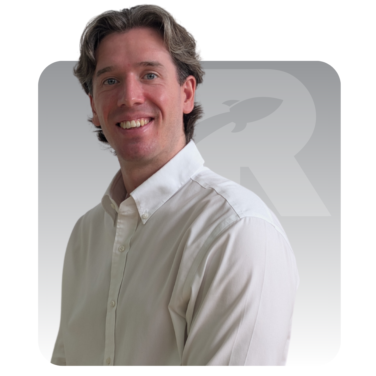 Smiling man in a white shirt standing against a white background with a large faint “R” logo.