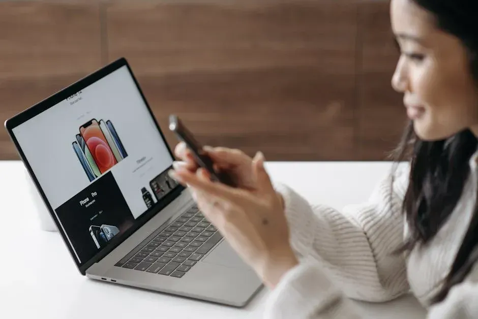 A woman is sitting at a table using a laptop and a cell phone.