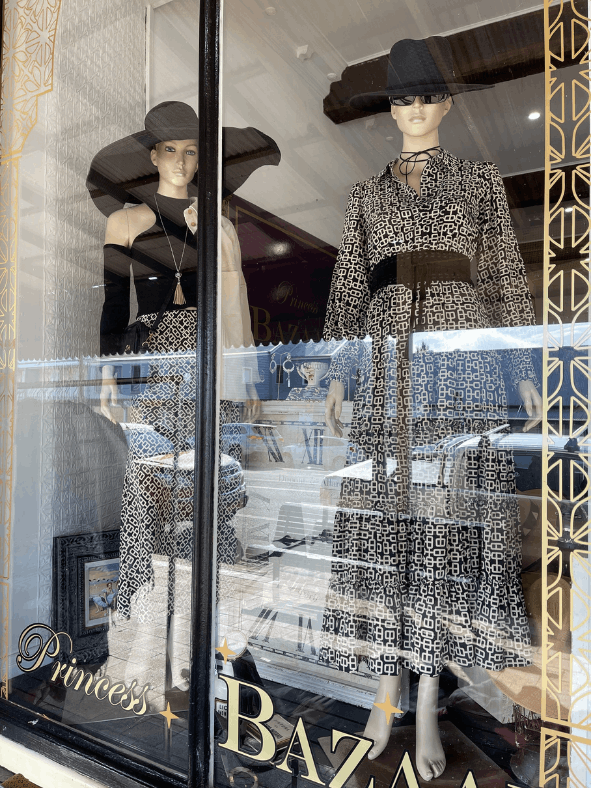 Two mannequins wearing hats and dresses are in a store window.