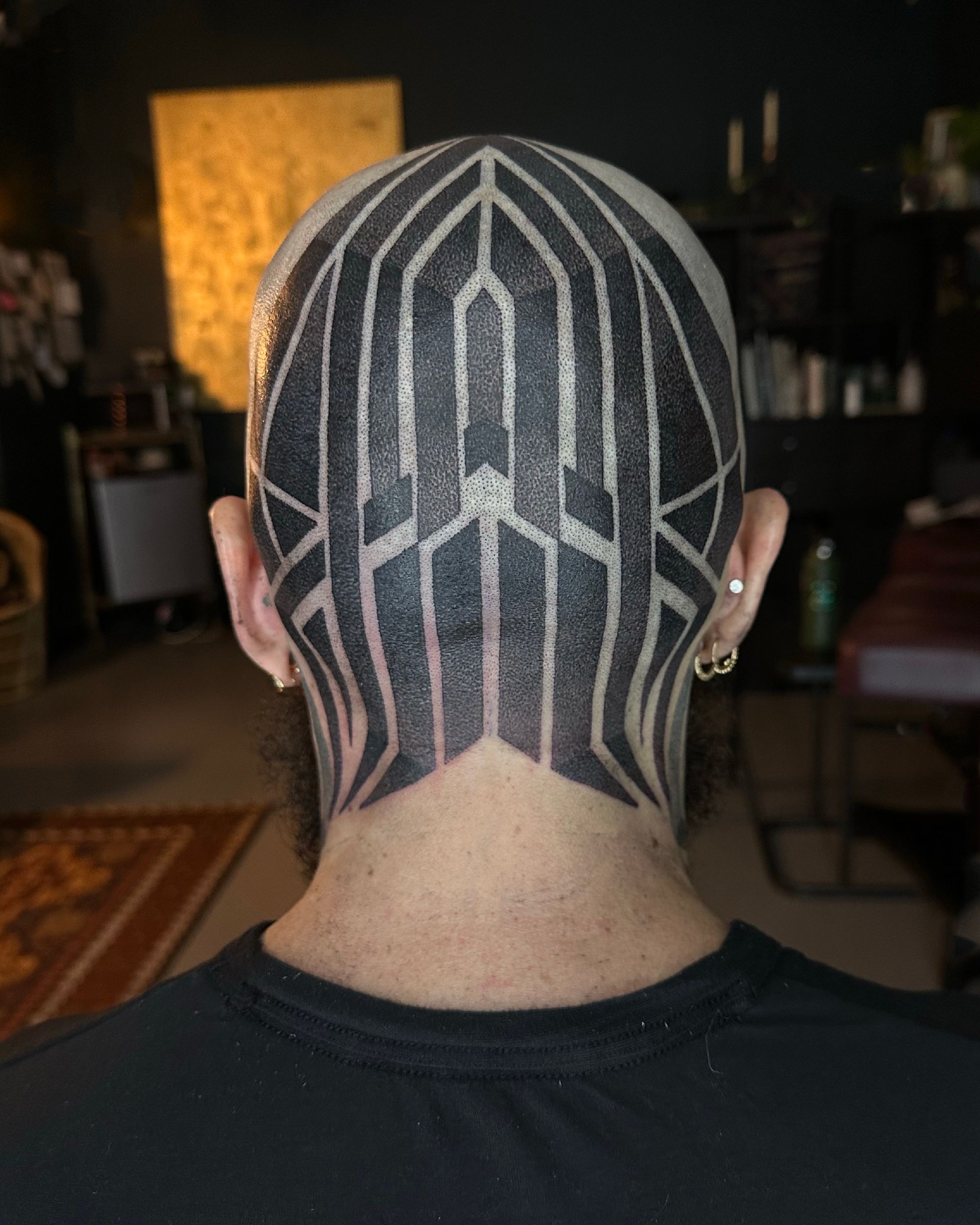 A person's shaved head with a geometric black tattoo pattern covering the back of their head and neck.