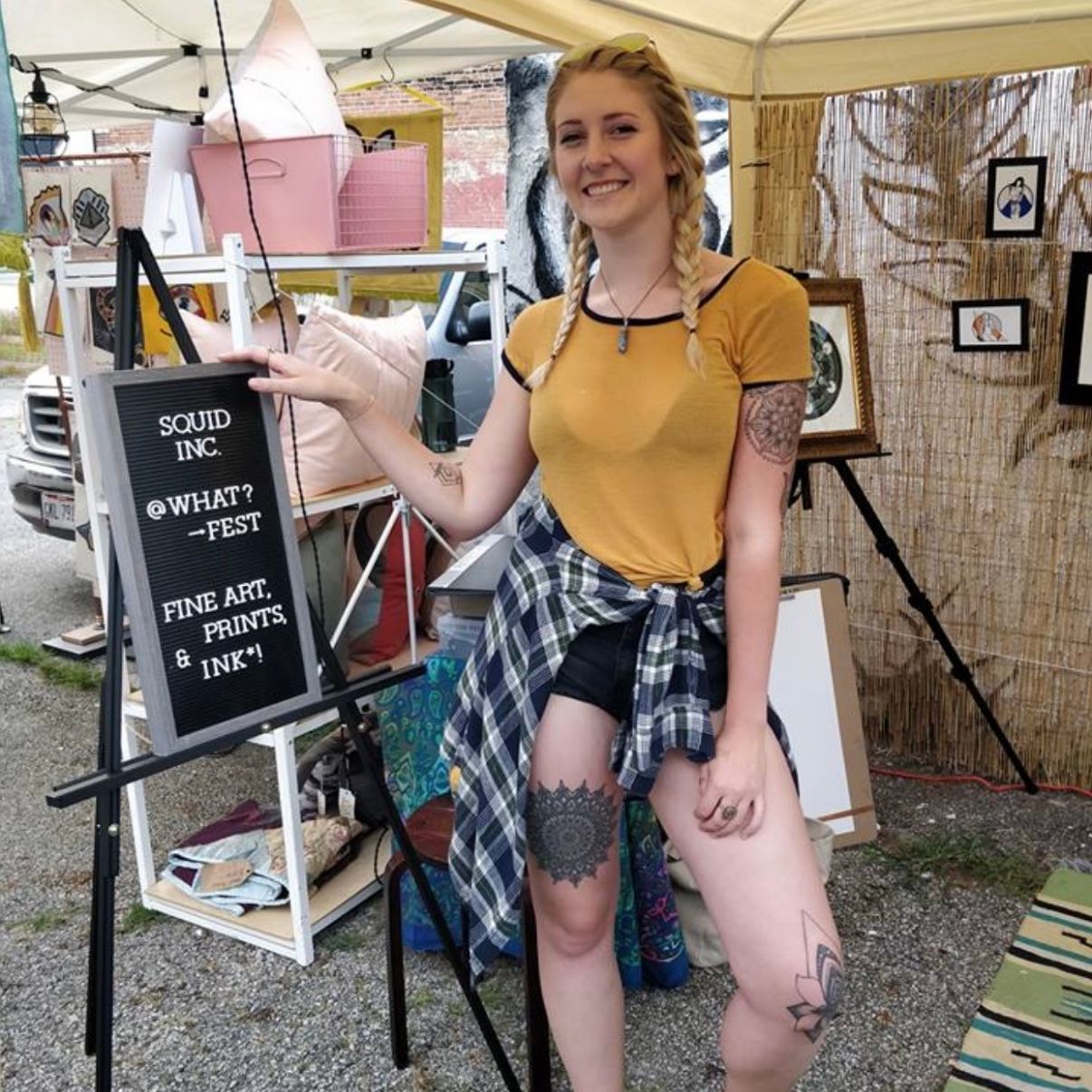 Woman at an outdoor craft fair, pointing to a sign. She wears a yellow shirt, shorts, and has tattoos; shop display behind.