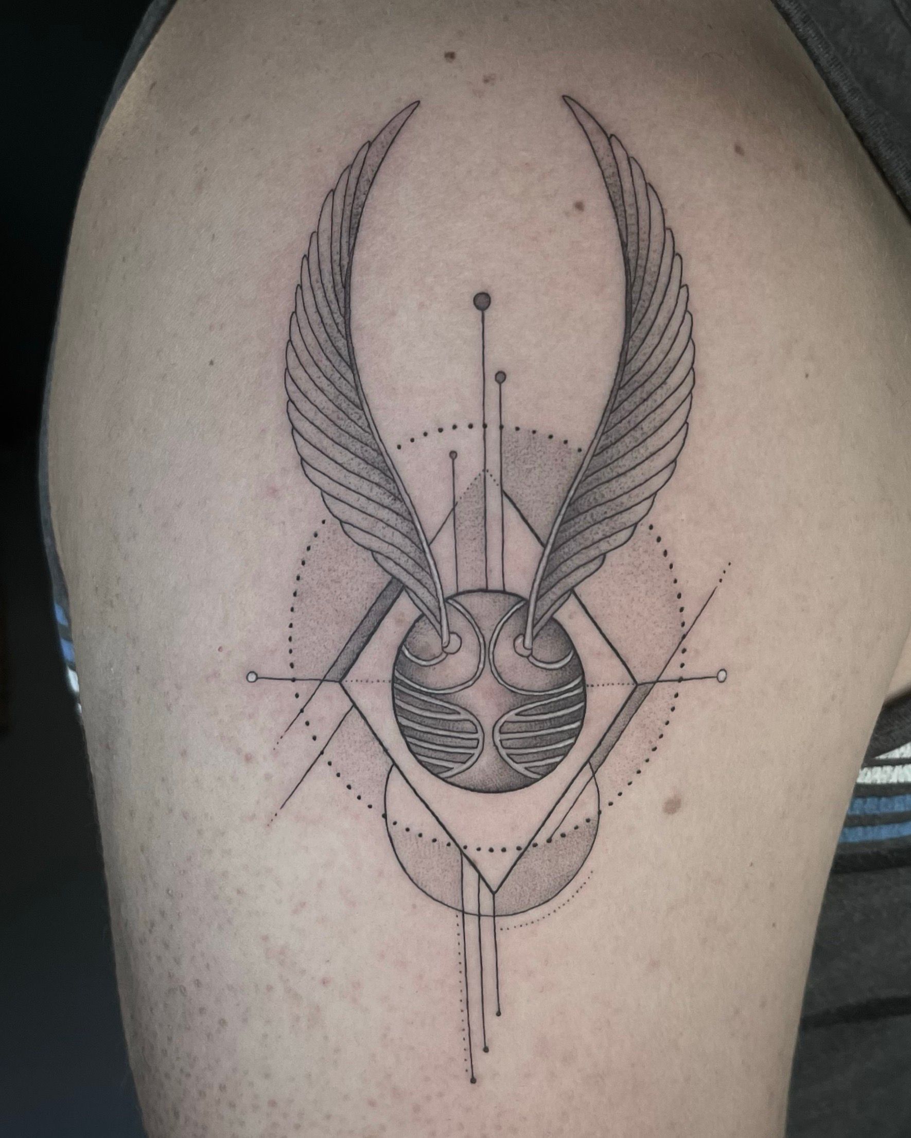 Tattoo of a golden snitch with wings and geometric lines on an upper arm.