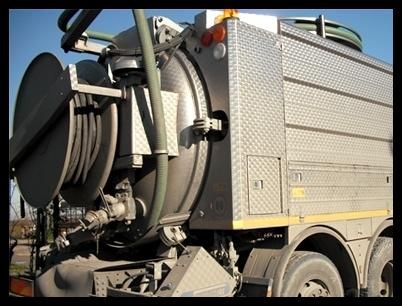Camion industriale grigio con grandi avvolgitubo e serbatoio metallico, parcheggiato all'aperto sotto il sole.