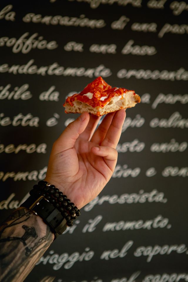 Una mano che regge una fetta di pizza contro una lavagna sfocata con testo italiano scritto a mano.