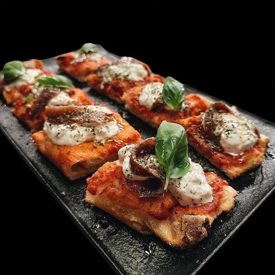Fette di pizza rettangolari carbonizzate, condite con formaggio fuso, salsa di pomodoro, basilico e olive, servite su un piatto nero.