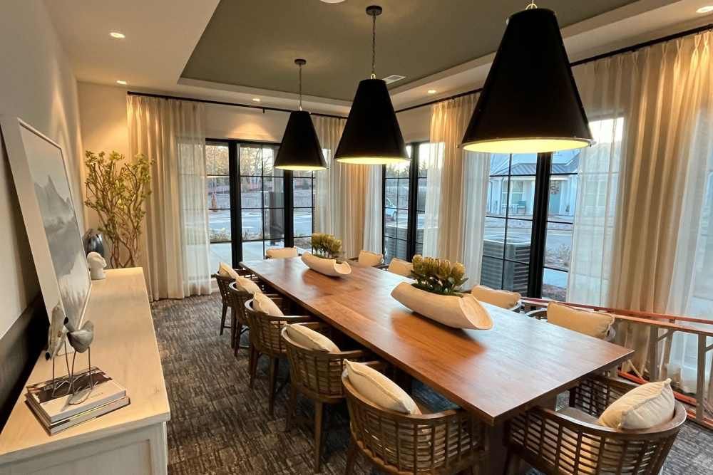 Dining room with long wooden table, black pendant lights, and woven chairs. Large windows, white curtains.