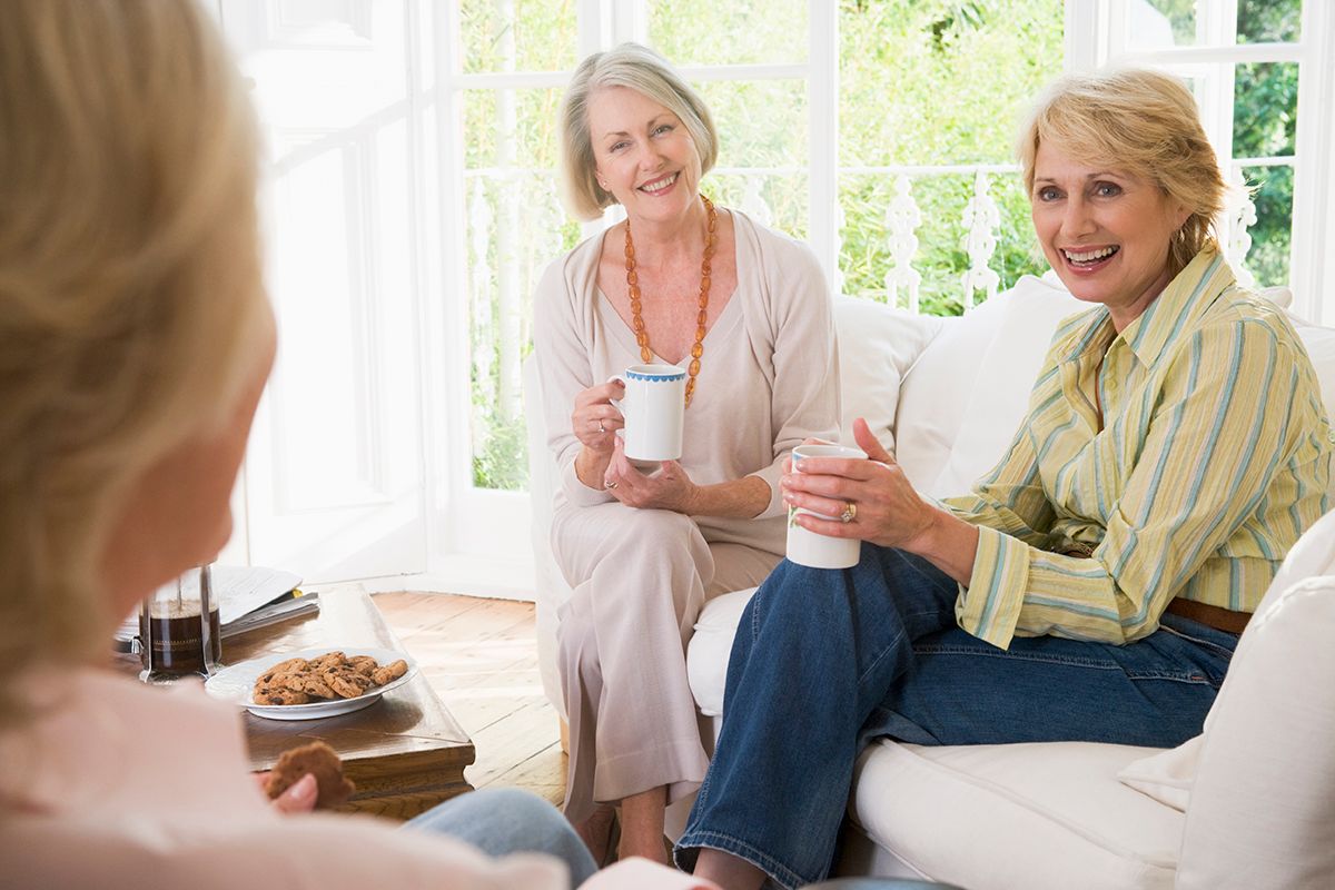 Three smiling women on a couch, holding mugs, enjoying a conversation in a well-lit living room.