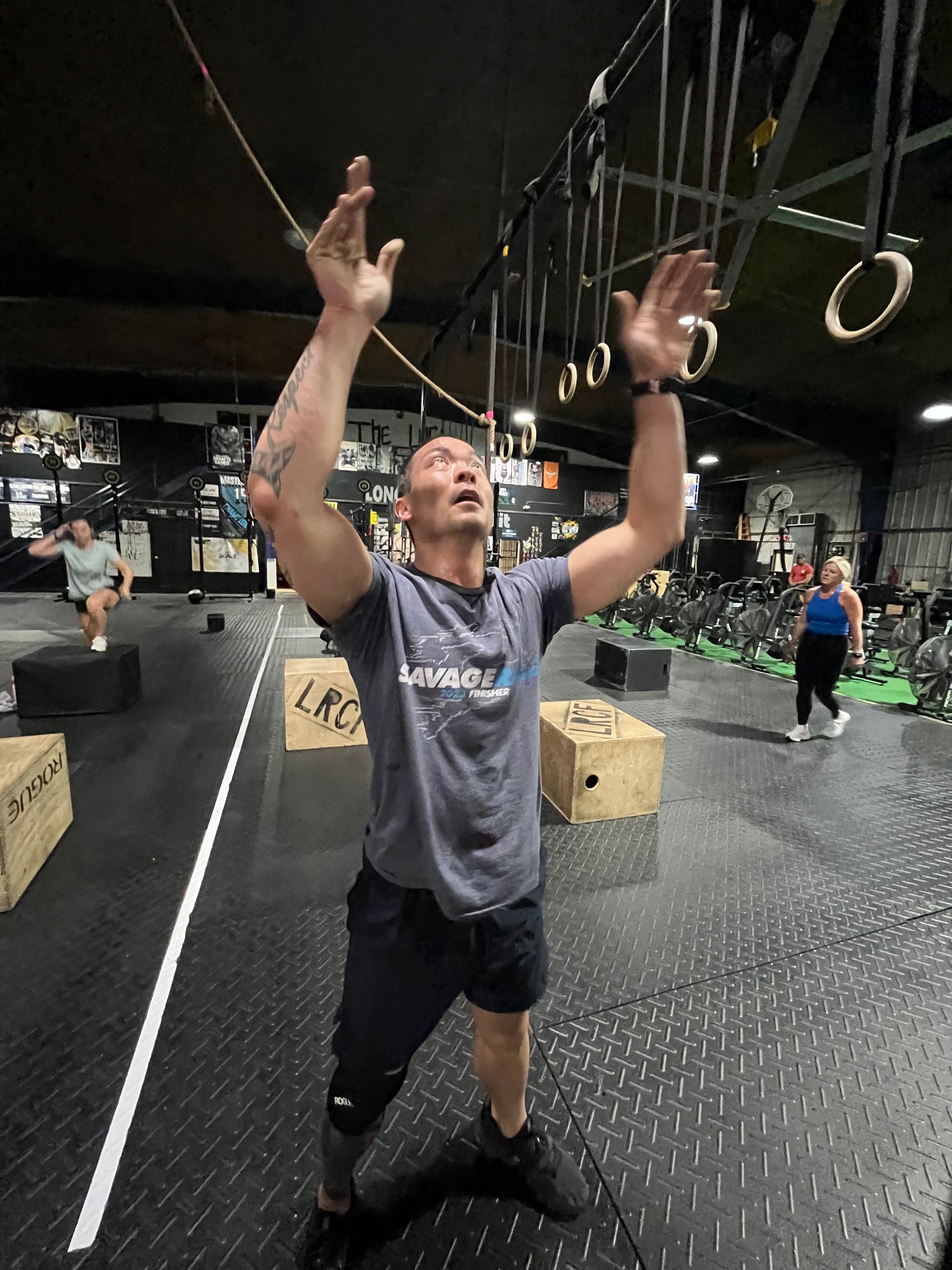 Person reaching up toward gymnastic rings in a CrossFit gym, surrounded by boxes and workout equipment.